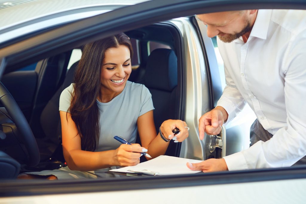 Eine lächelnde Frau sitzt im Auto und unterschreibt den Kaufvertrag, den ihr ein Autohändler hinhält.