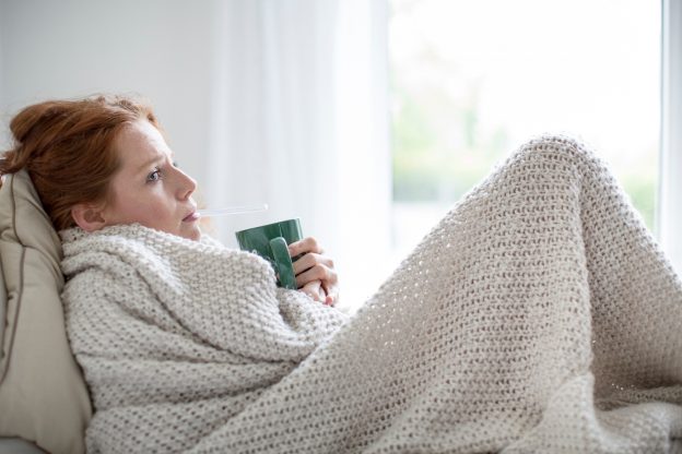 Kranke Frau sitzt in eine Decke gehüllt und hält eine Teetasse.