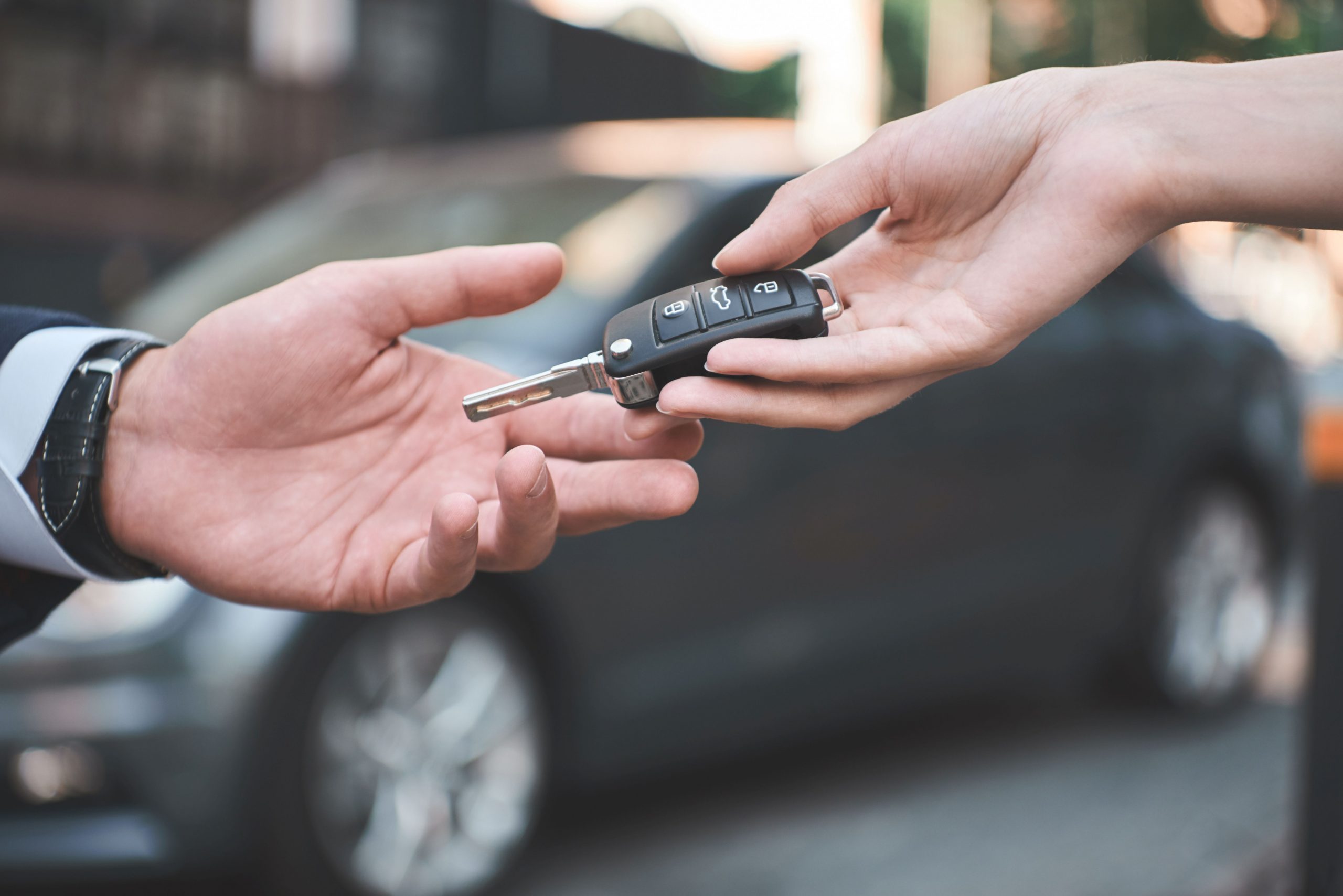 Eine Person überreicht einer anderen Person einen Autoschlüssel. Im Hintergrund steht ein dunkelgraues Auto.